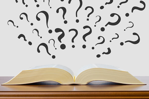 An opened book with question marks flying out of the pages resting on a wooden table.