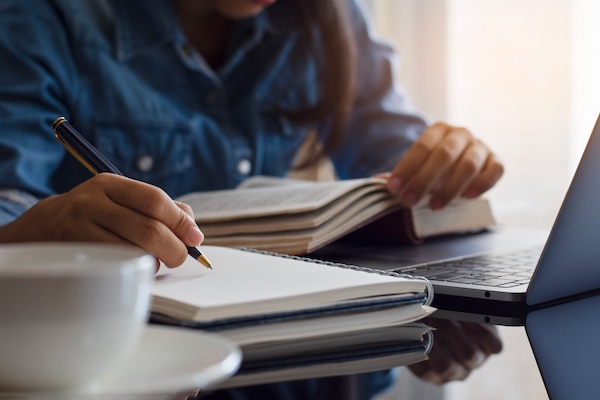 Casual young female student handwriting information on diary notebook while reading book and work on laptop computer with white cup of coffee on the table at home.