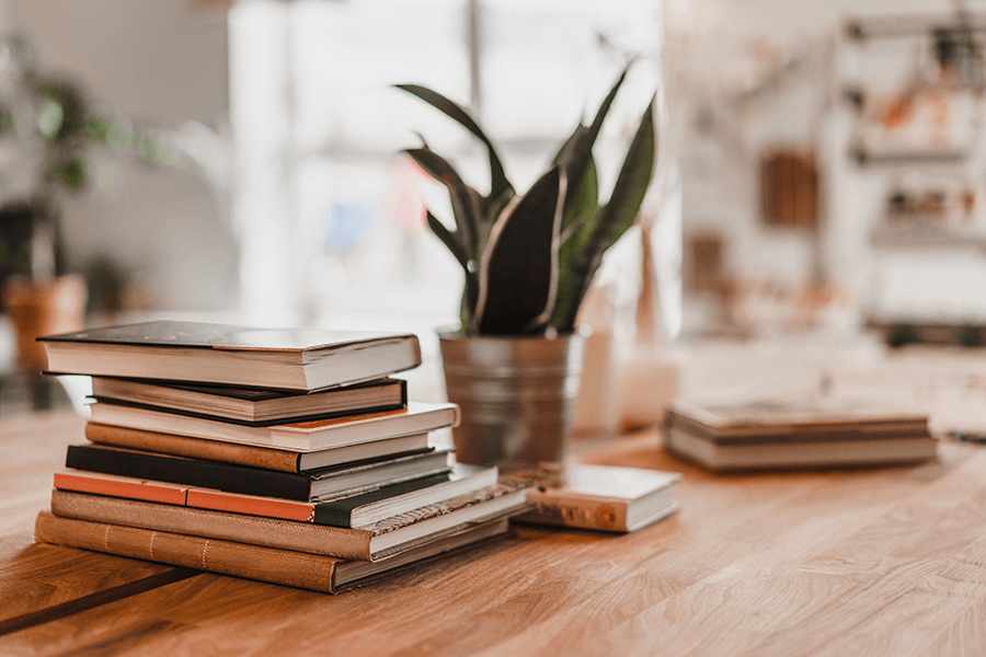 Stack of colorful books on wooden table in a home office next to a house plant