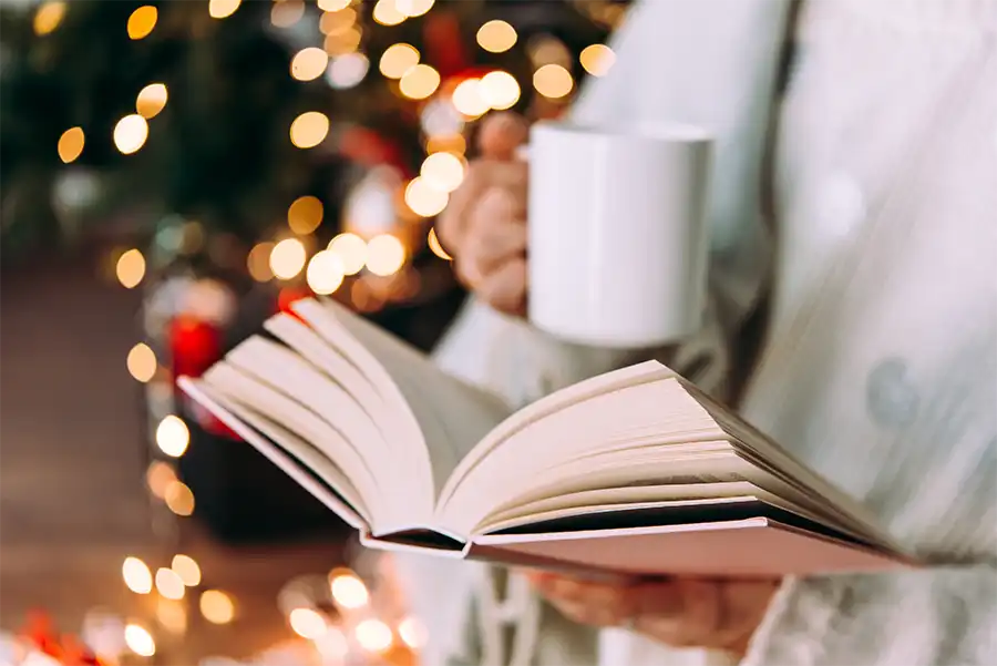 Dealing with Loneliness as a Writer During the Holidays- Finding Connection and Joy close up image of a woman's hands. One is holding an open book and her other hand is holding a white coffee mug. There are Christmas lights behind her, but they are out of focus